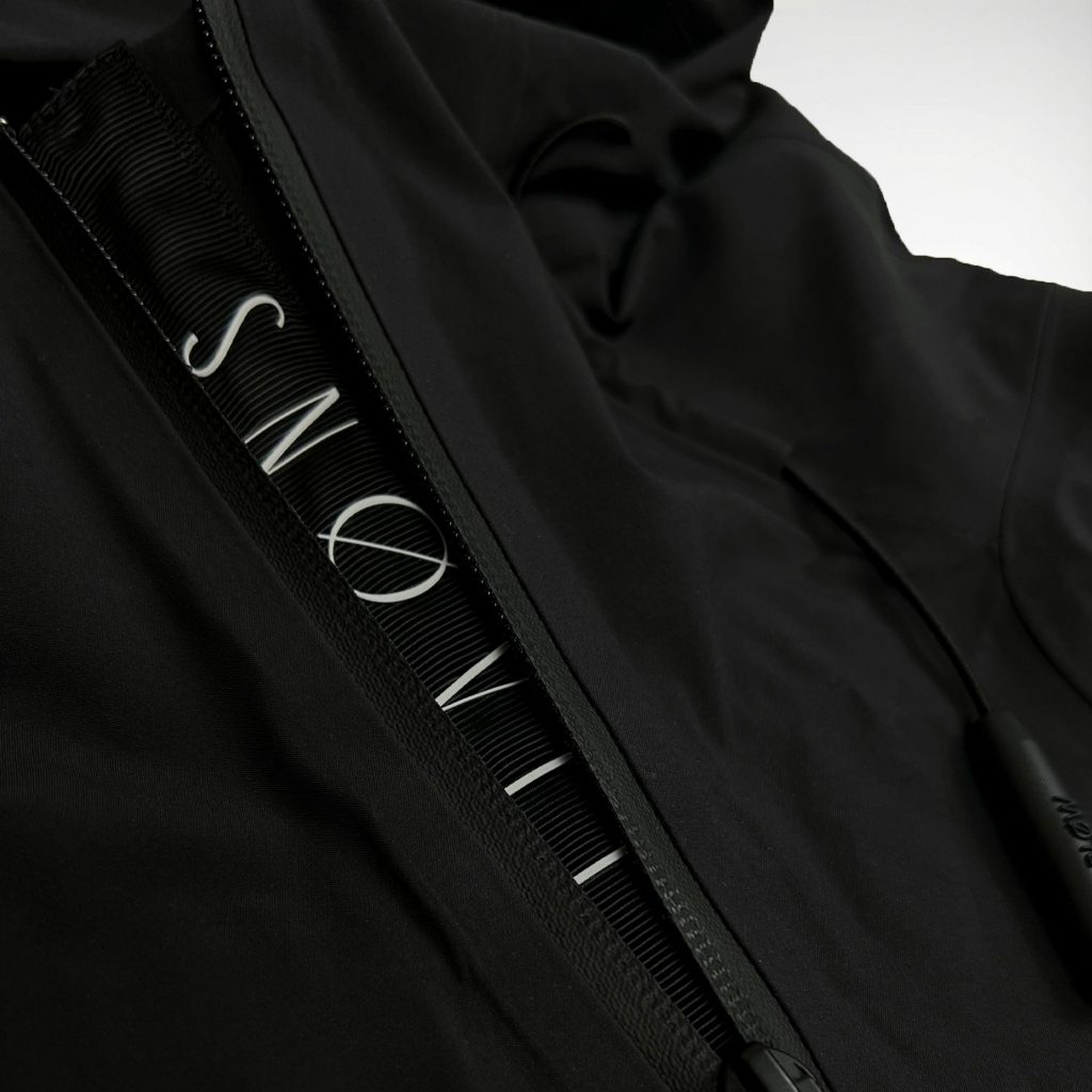 Nahaufnahme des gewebten SNØVIT Stripes an der schwarzen Nithya Nøva X Outdoor-Jacke, hochwertige Nylonstruktur sichtbar.