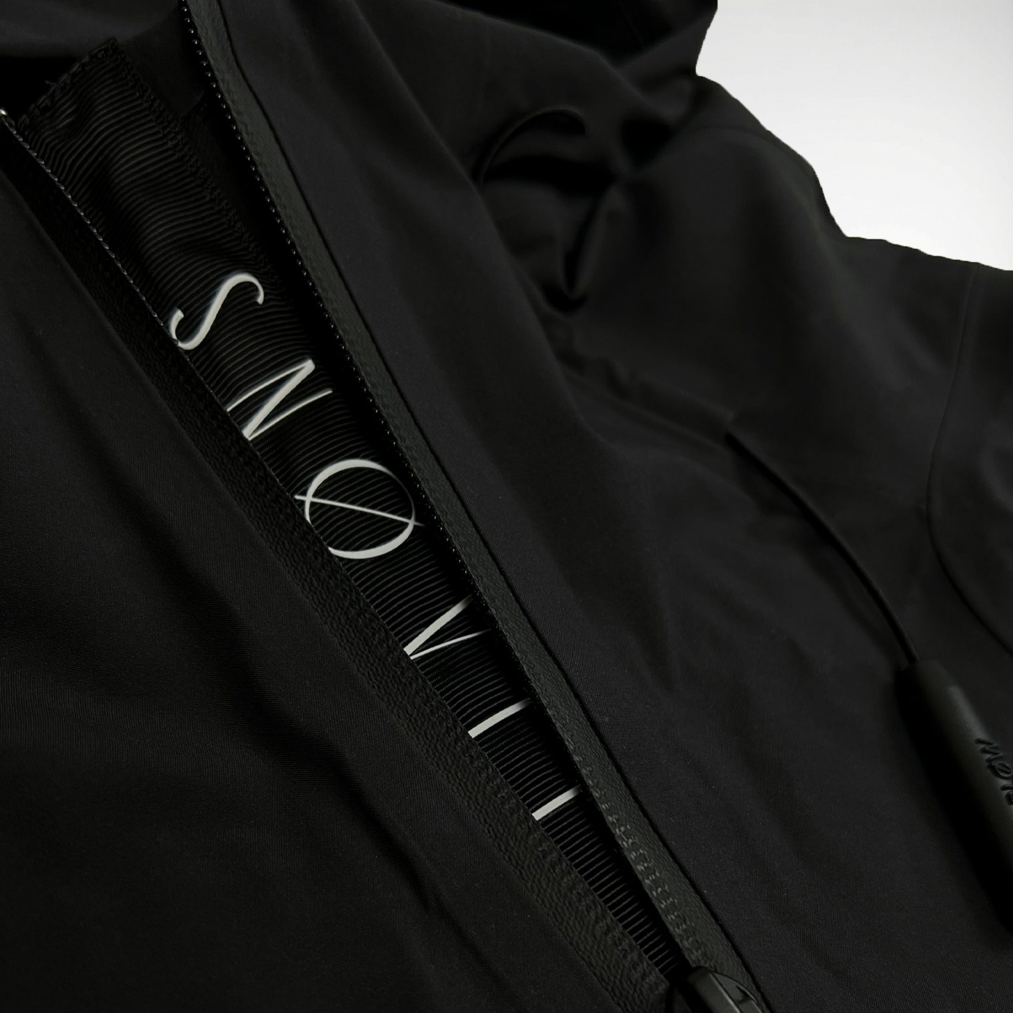 Nahaufnahme des gewebten SNØVIT Stripes an der schwarzen Nithya Nøva X Outdoor-Jacke, hochwertige Nylonstruktur sichtbar.