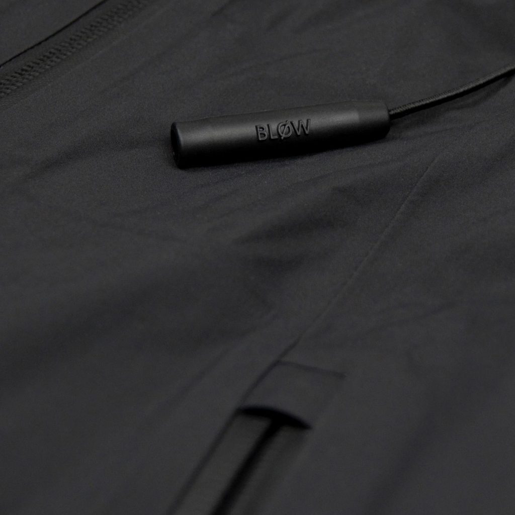 Nahaufnahme der eingebauten Notfall-Pfeife „BLØW“ an der SNØVIT Nithya Nøva X Outdoor-Jacke, inspiriert vom Alpengürtelmurmeltier.