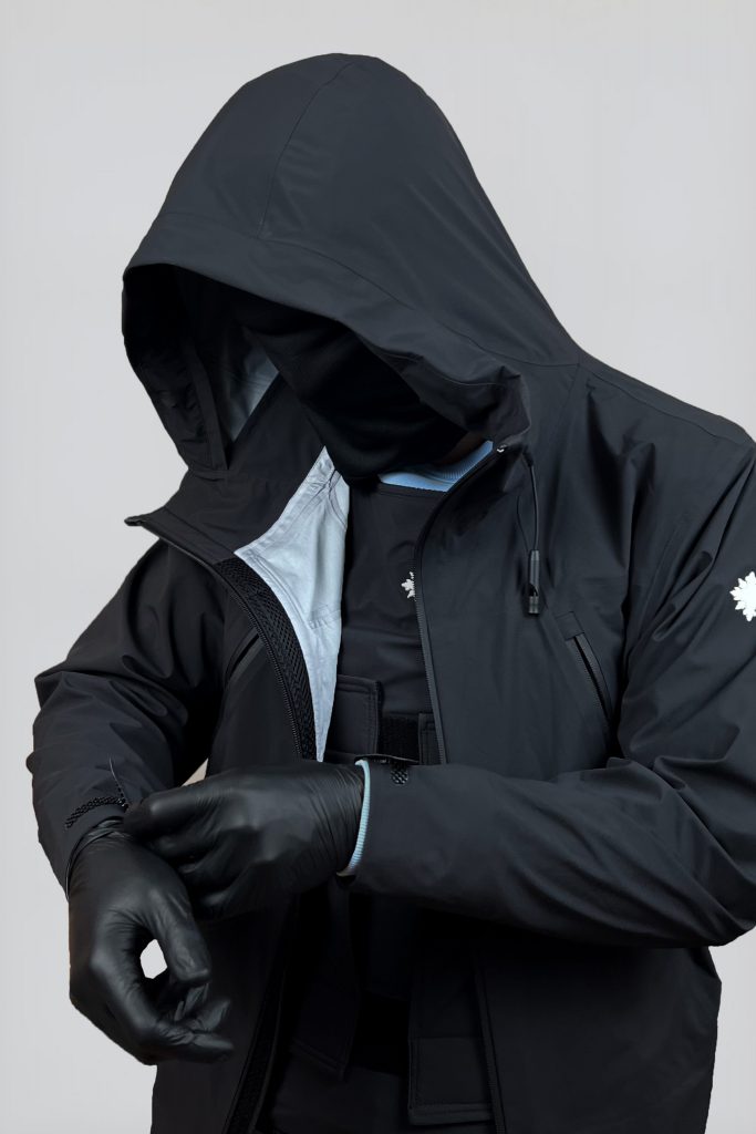 Hochqualitatives Bild eines Models, das die schwarze SNØVIT Nithya Nøva X Outdoor-Jacke trägt, mit der diskret darunter getragenen DEFENX PRO Vanta Weste.