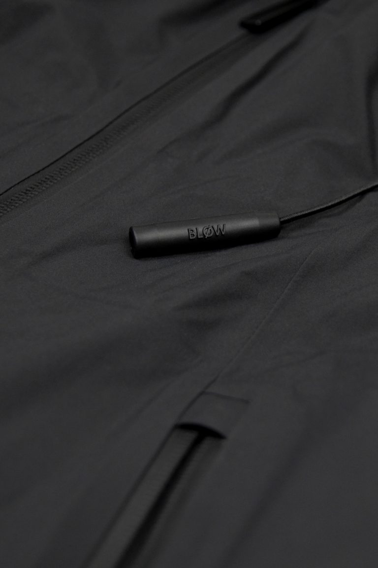 Nahaufnahme der eingebauten Notfall-Pfeife „BLØW“ an der SNØVIT Nithya Nøva X Outdoor-Jacke, inspiriert vom Alpengürtelmurmeltier.