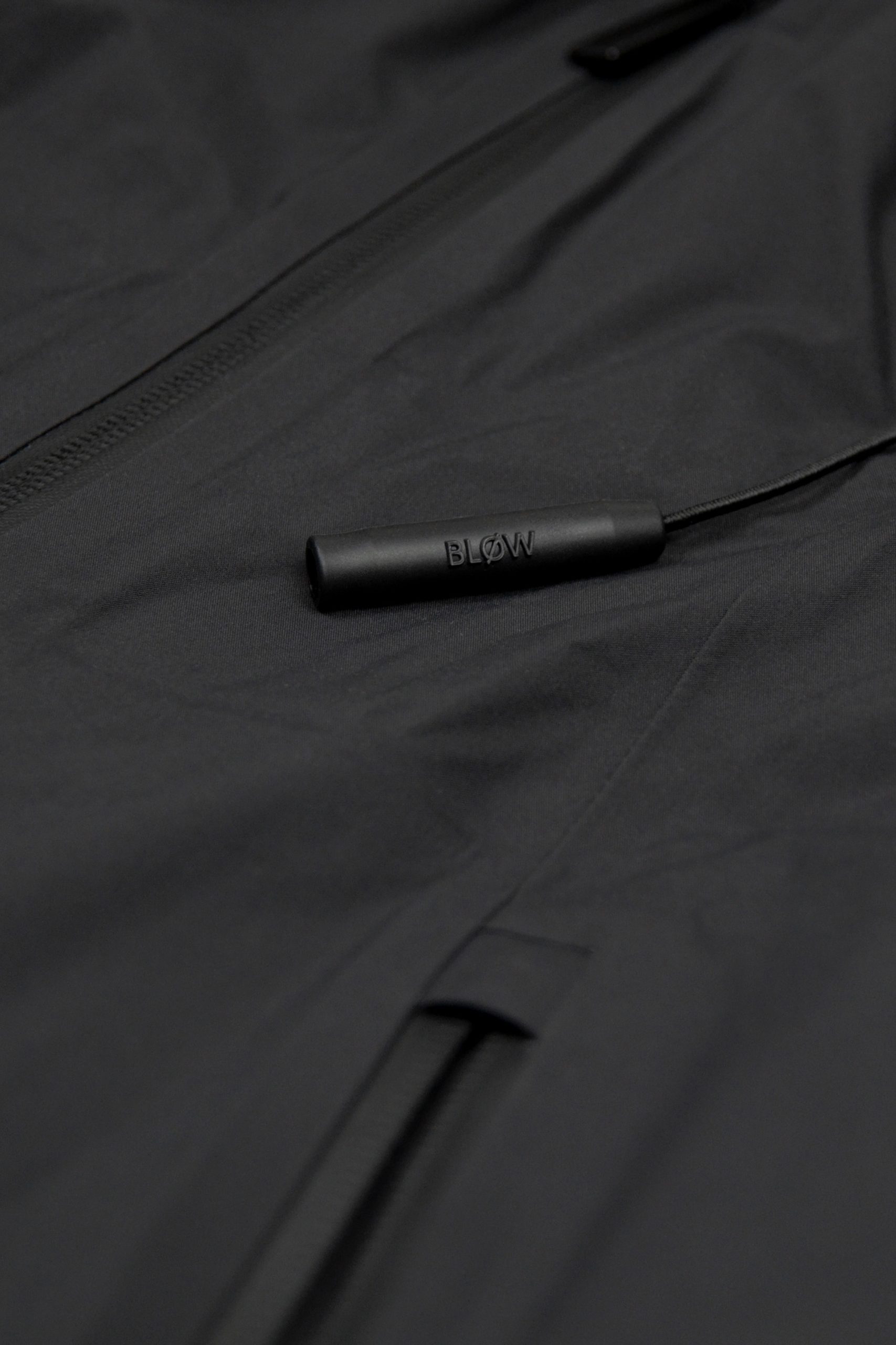 Nahaufnahme der eingebauten Notfall-Pfeife „BLØW“ an der SNØVIT Nithya Nøva X Outdoor-Jacke, inspiriert vom Alpengürtelmurmeltier.